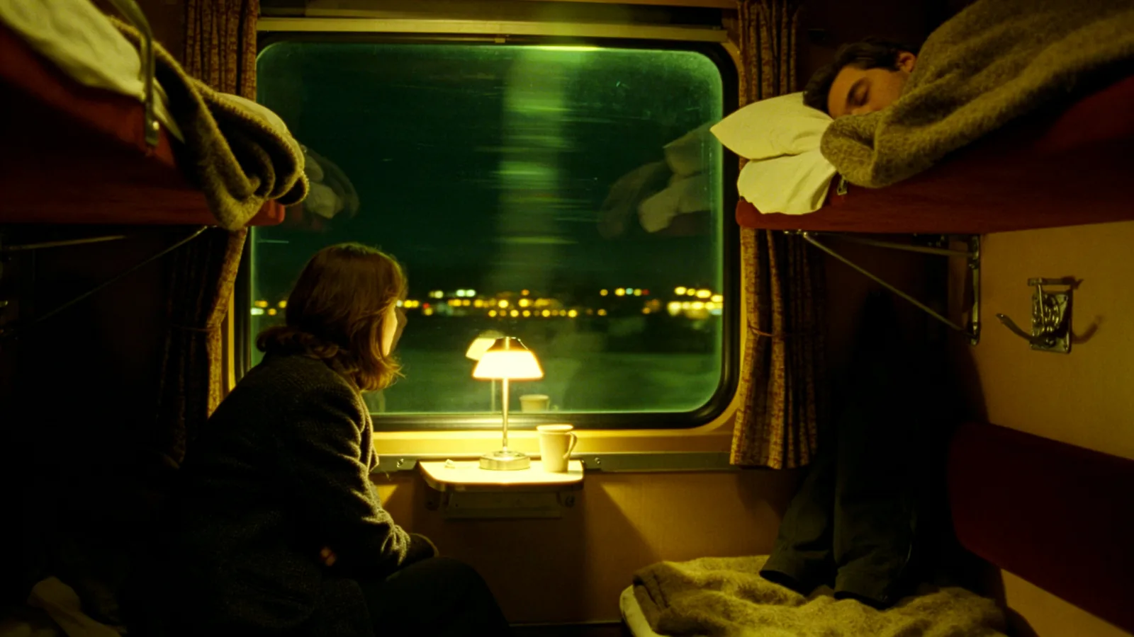 Dos viajeros en un compartimento con literas de un tren nocturno; uno mira pasar las luces por la ventana.